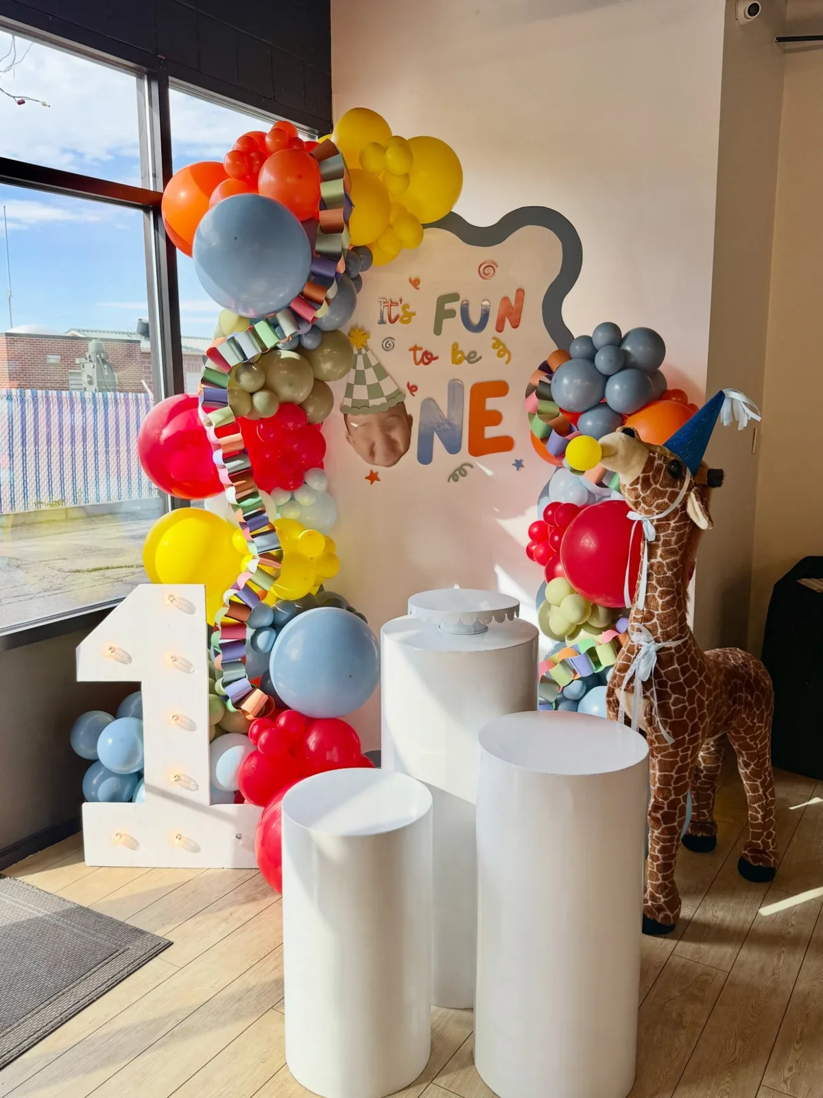 A colorful first birthday balloon garland with a number 1 marquee, round pedestals, and a giraffe plush