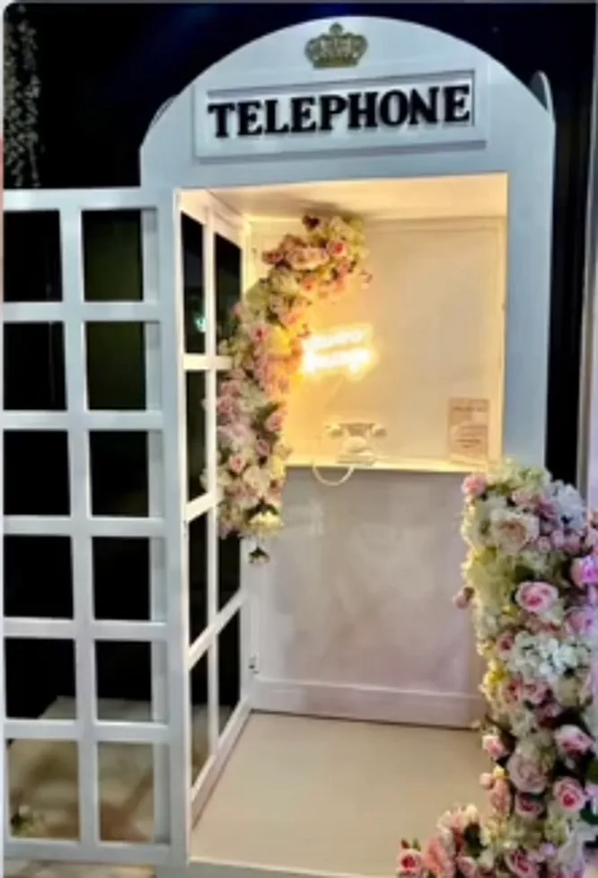A white vintage telephone booth with a Telephone sign, a neon light, and pink and white flowers