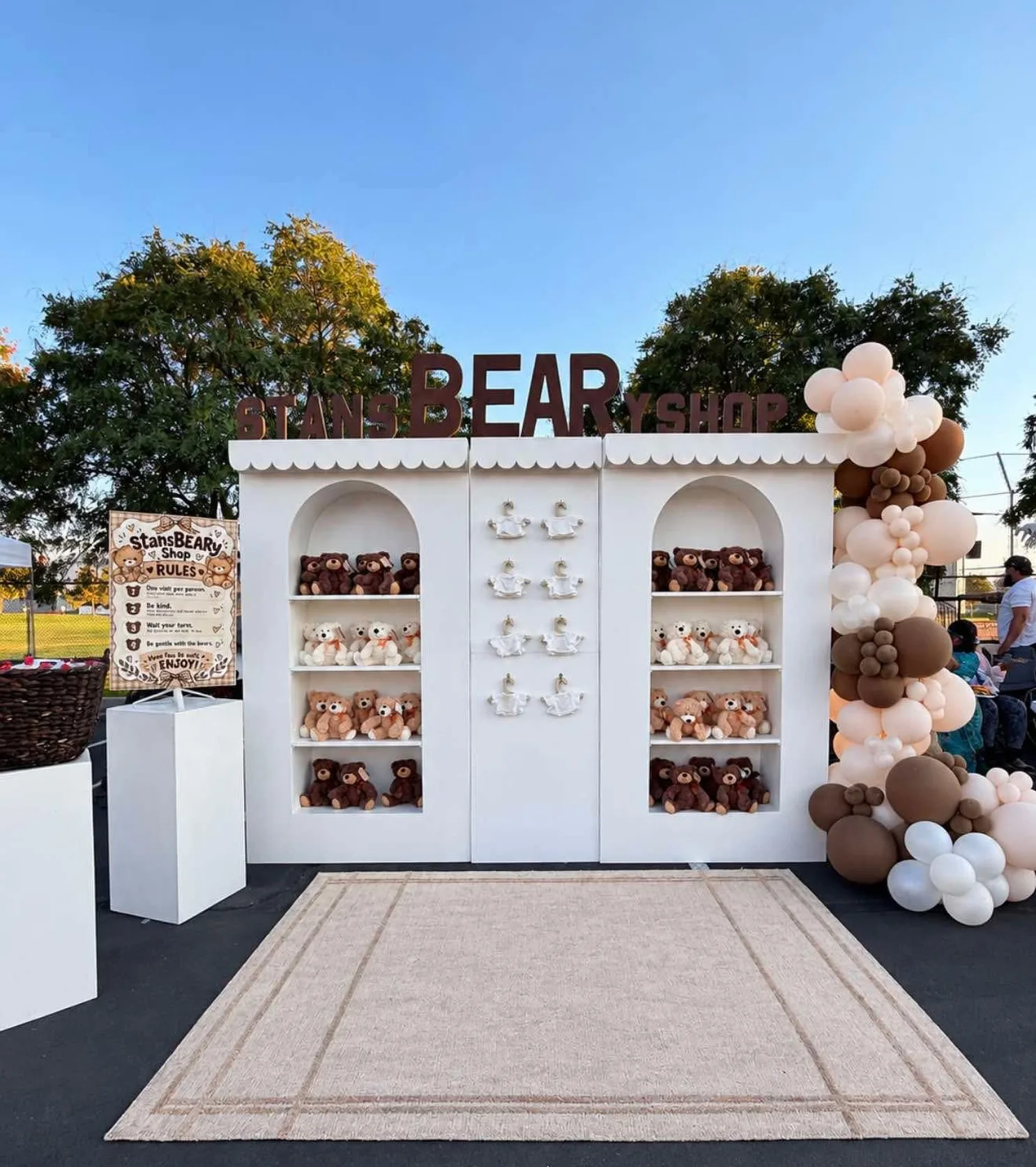 A white pop-up teddy bear shop with arched shelves, a scalloped roof, and a brown and cream balloon garland