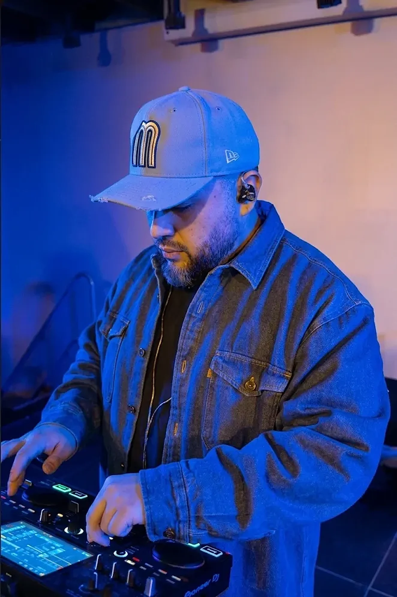 DJ Iguana in a denim jacket and a cap, mixing on a DJ controller under blue light