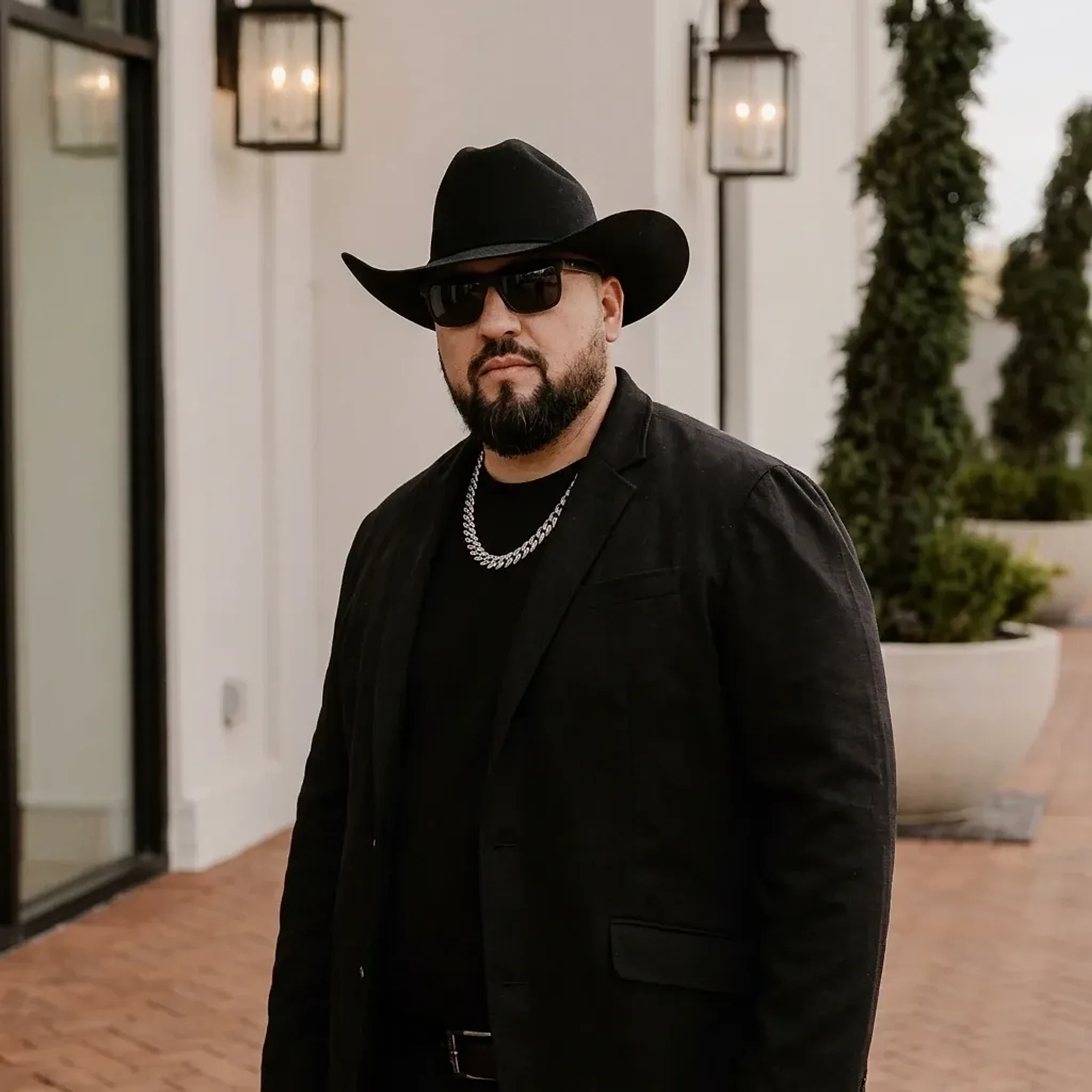DJ Iguana in a black cowboy hat, sunglasses, and a black jacket outside a white building