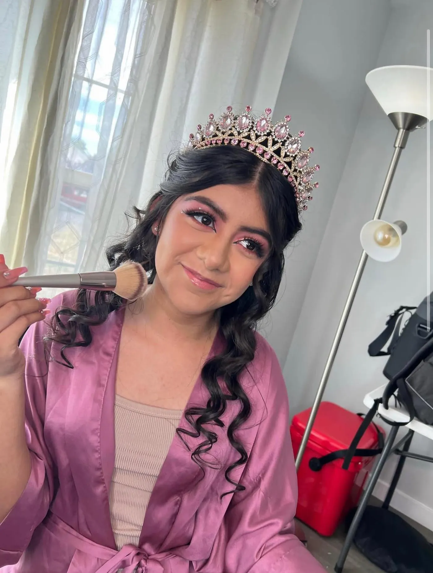 A quinceañera in a pink robe and a pink tiara having her makeup finished