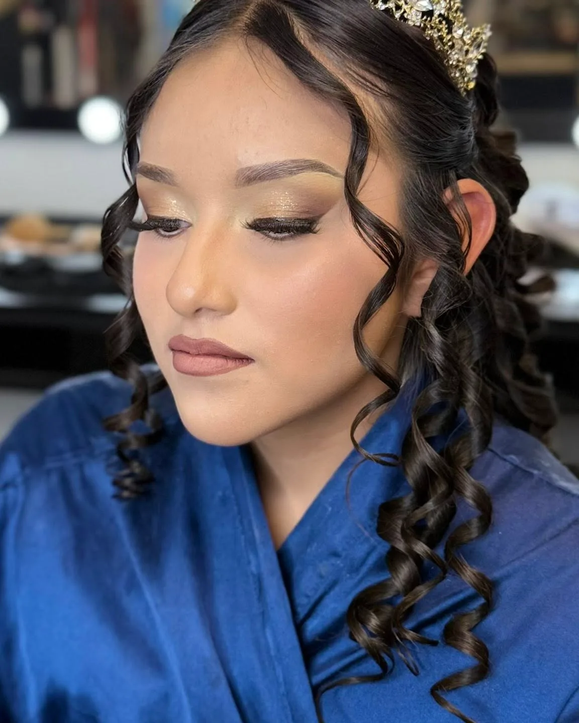 A client with golden shimmer eyeshadow, a winged liner, and long curls beneath a crown