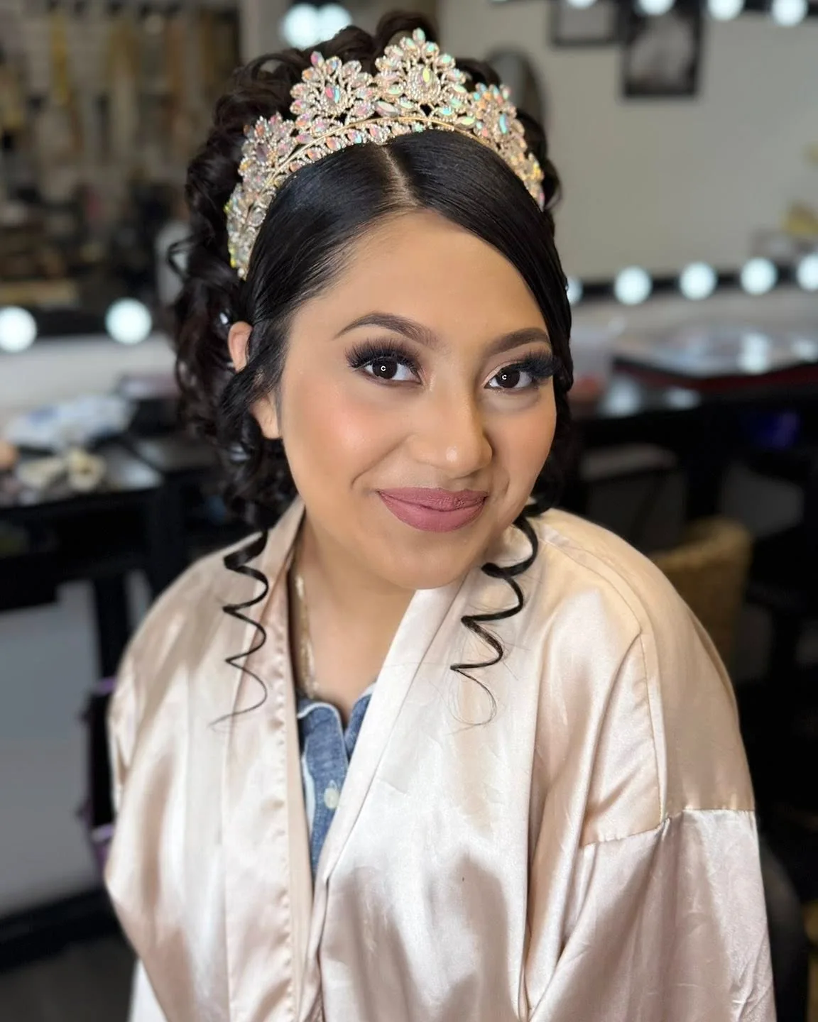 A quinceañera in a pearl-colored robe with soft glam makeup, curls, and a crystal crown