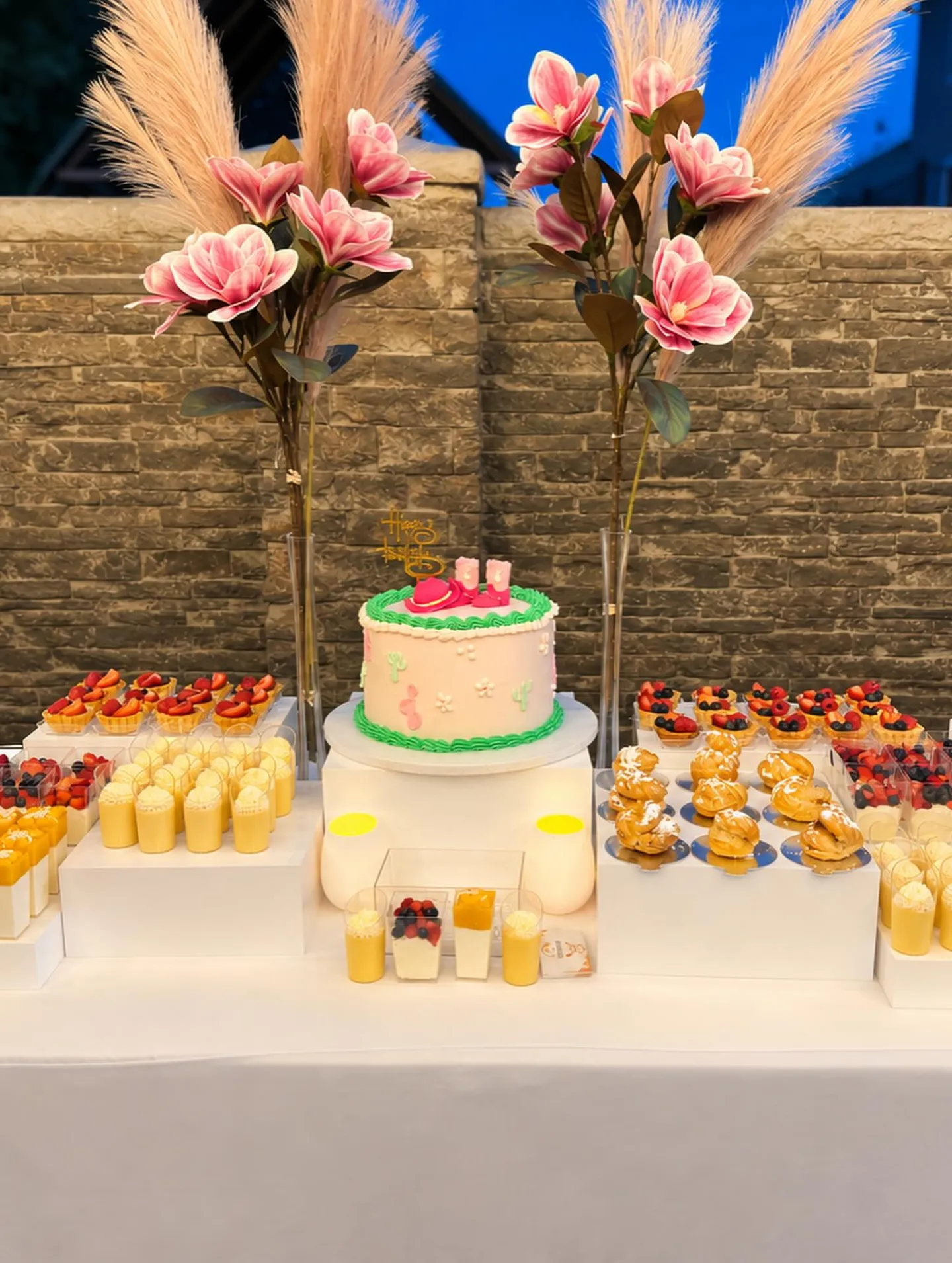 A dessert table with a pink cake between pampas grass and magnolias