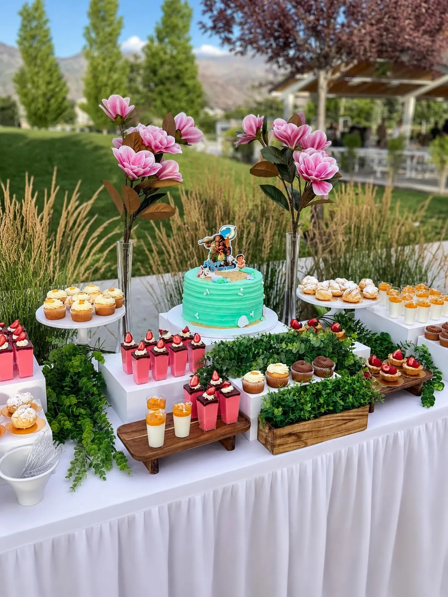 A Mini Bites dessert table outdoors with a teal cake, mousse cups, cupcakes, and pink magnolias