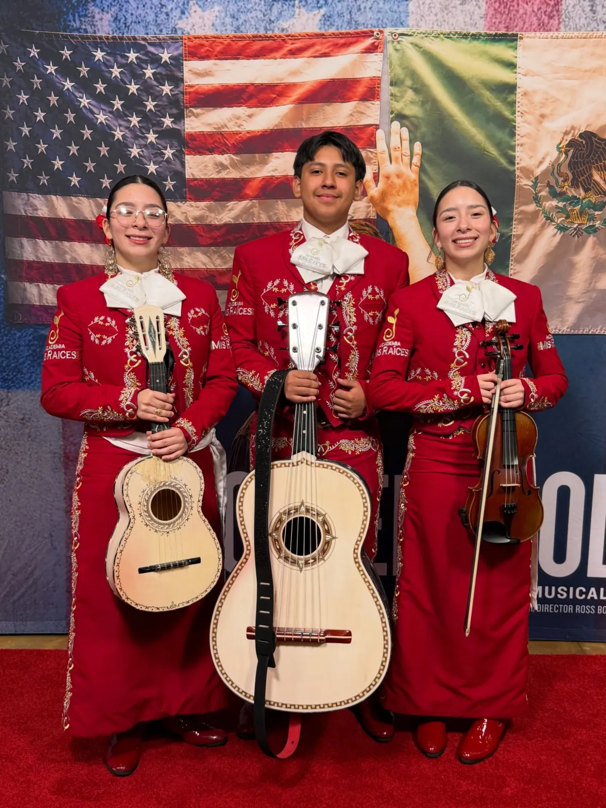 Three Academia Mis Raíces students in red mariachi trajes holding a vihuela, a guitarrón, and a violin