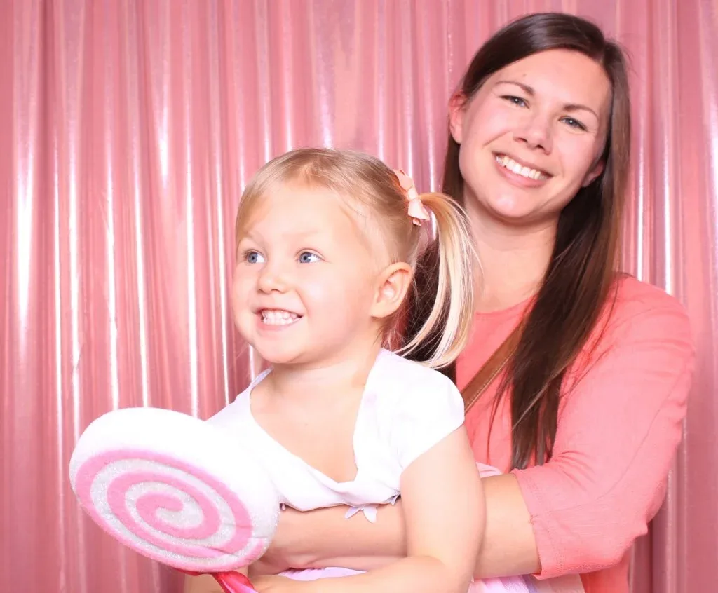 A mom and daughter with an oversized lollipop against pink shimmer