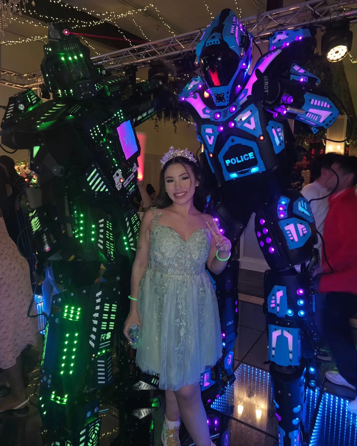 A quinceañera in a mint dress and crown posing between two LED robots