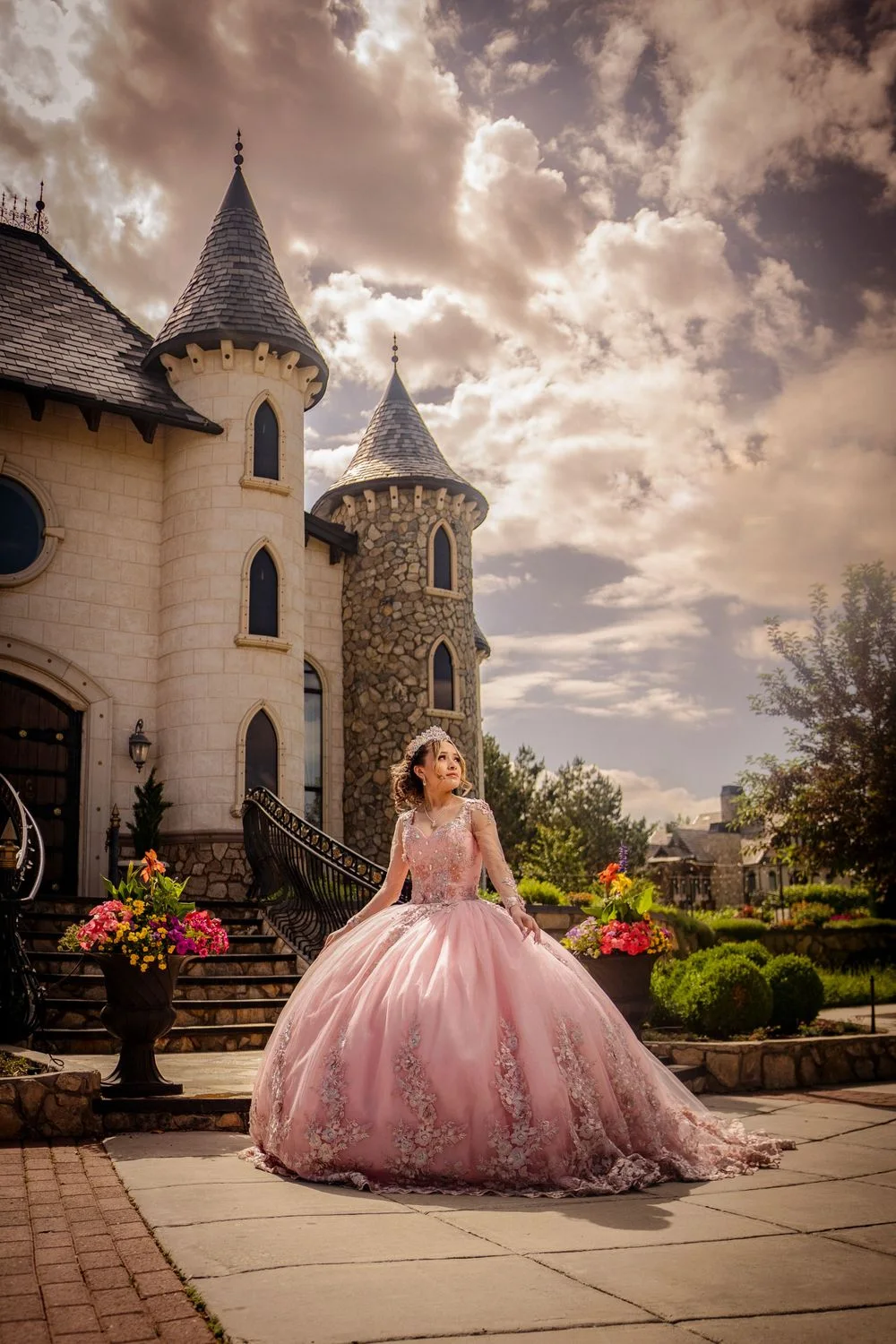 A quinceañera in a pink lace ball gown in front of a castle under a dramatic sky