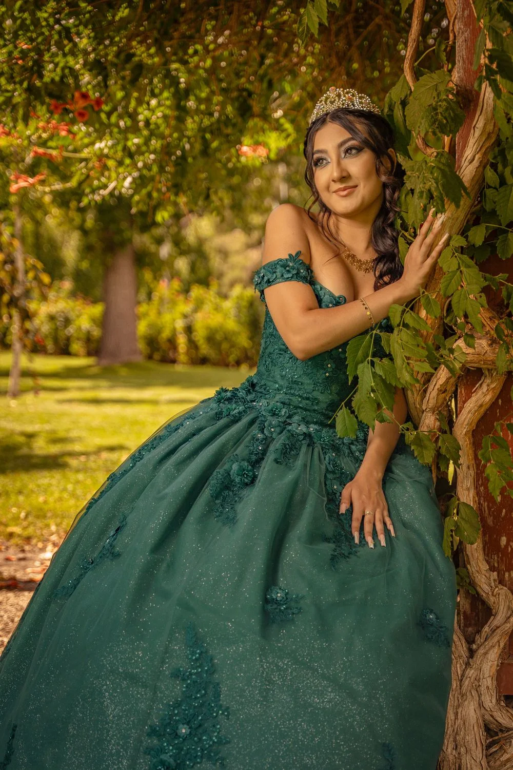 A young woman in an emerald off-shoulder gown leaning against a vine-covered tree