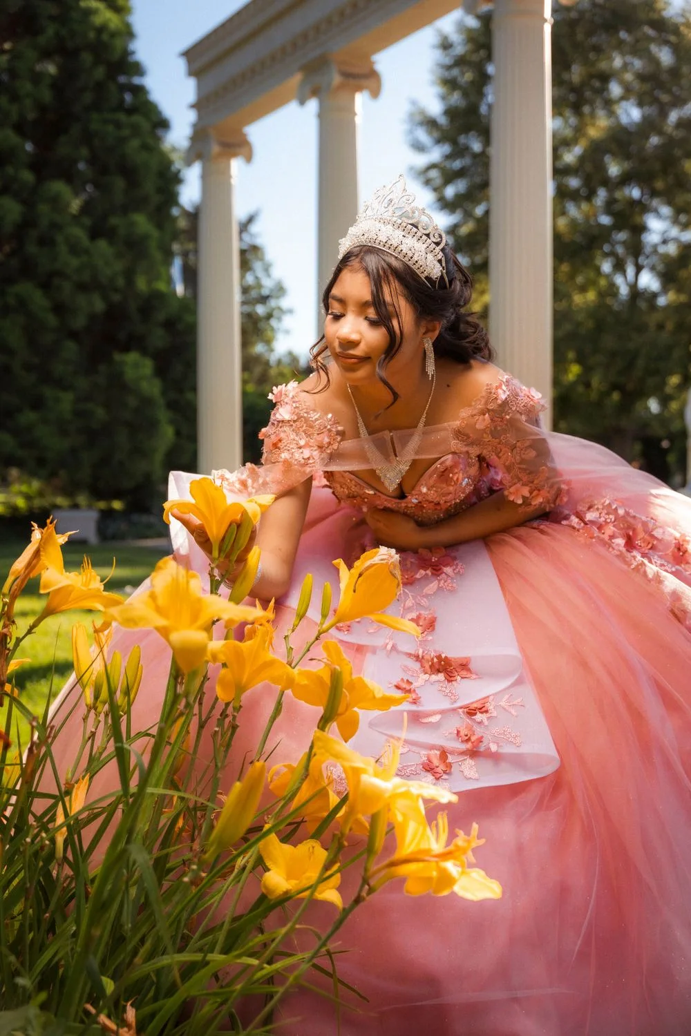 A young woman in a pink floral ball gown and tiara admiring yellow lilies in a garden