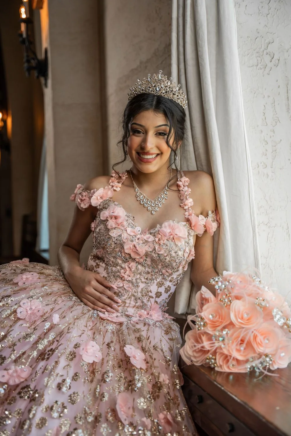 A smiling quinceañera in a pink floral gown with a tiara, holding a rose bouquet