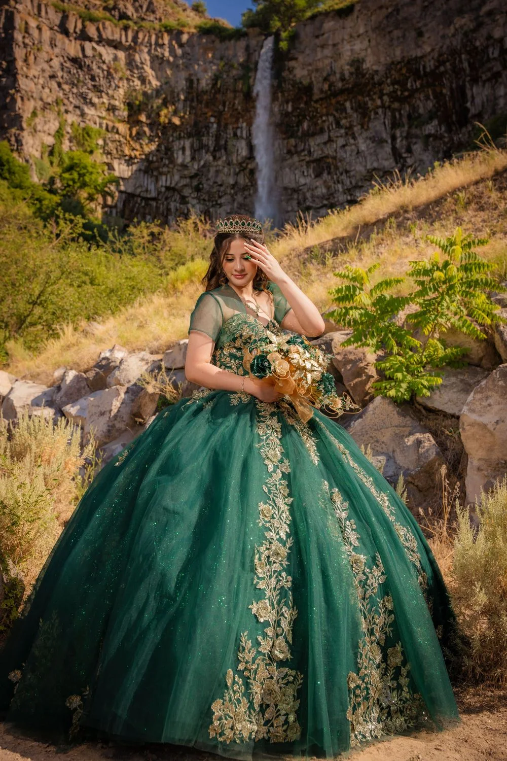 A young woman in an emerald green quinceañera gown holding a bouquet near a waterfall