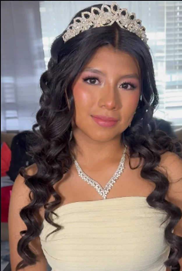 A quinceañera with long dark curls, pink eyeshadow, a tiara, and a crystal necklace